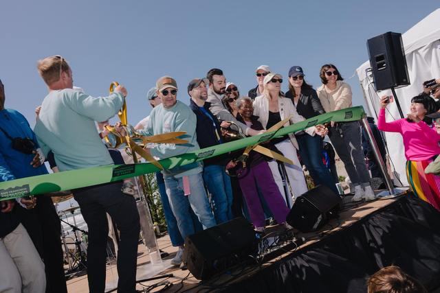 A group of people stands on a stage, cutting a green ribbon with oversized scissors during an outdoor event. A crowd watches, and someone takes a photo.